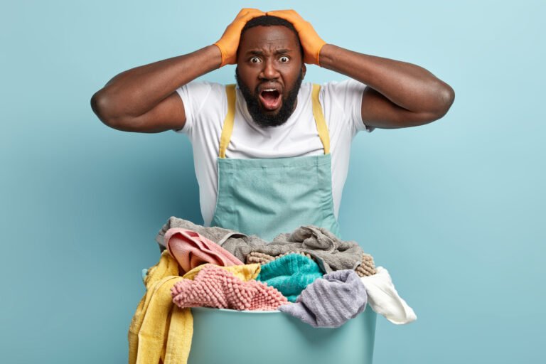 Frustrated dark skinned bearded man keeps both hands on head, face problem of brocken washing machine, has to do hand wash, busy with household duties, laundry, stands near basin of dirty clothes